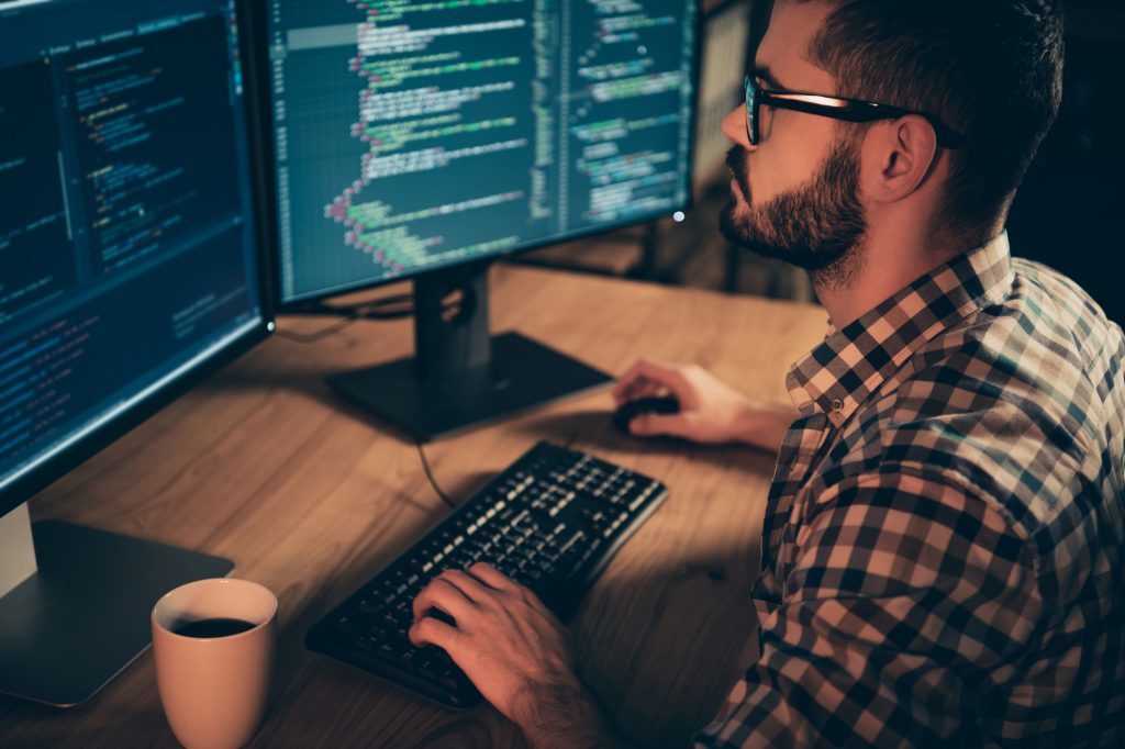 Close up side profile photo handsome he him his guy linux windows cross platform coder typing php css keyboard development outsource IT monitors table office agency wear specs formalwear plaid shirt