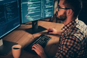 Close up side profile photo handsome he him his guy linux windows cross platform coder typing php css keyboard development outsource IT monitors table office agency wear specs formalwear plaid shirt