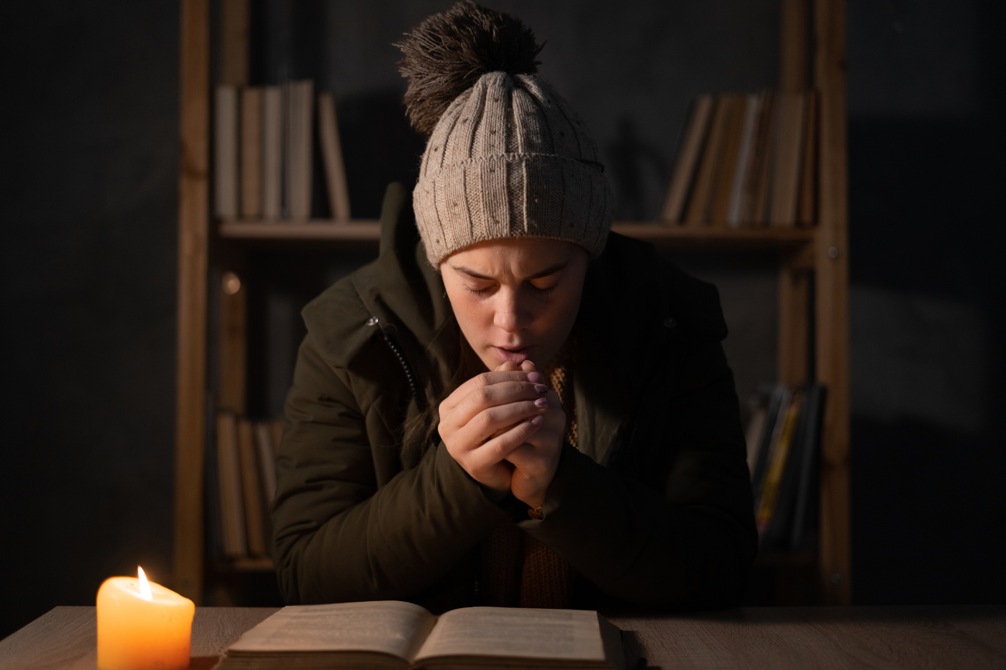 Freezing young woman in winter clothes warms her hands on lights with candles. Shutdown of heating and electricity, power outage, blackout, load shedding or energy crisis.