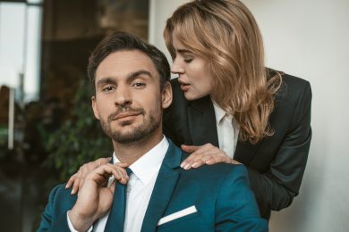 Passionate couple flirts at work. Office romance concept. Caucasian man and woman in love wearing formalwear. Man looks at camera, while woman whispers in his ear