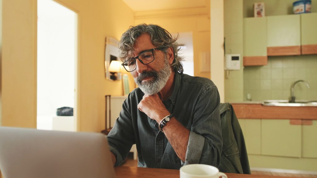 Elderly positive man typing on laptop sitting in home kitchen
