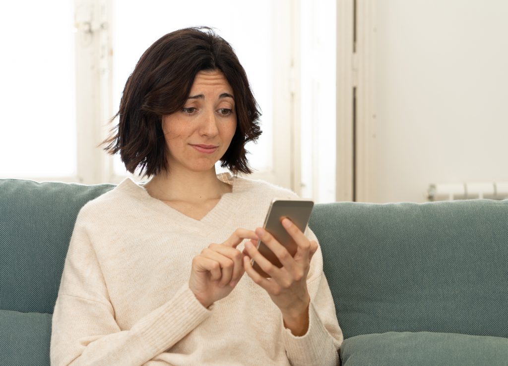 Happy young woman using her smart phone sitting on sofa at home. In leisure and mobile addiction