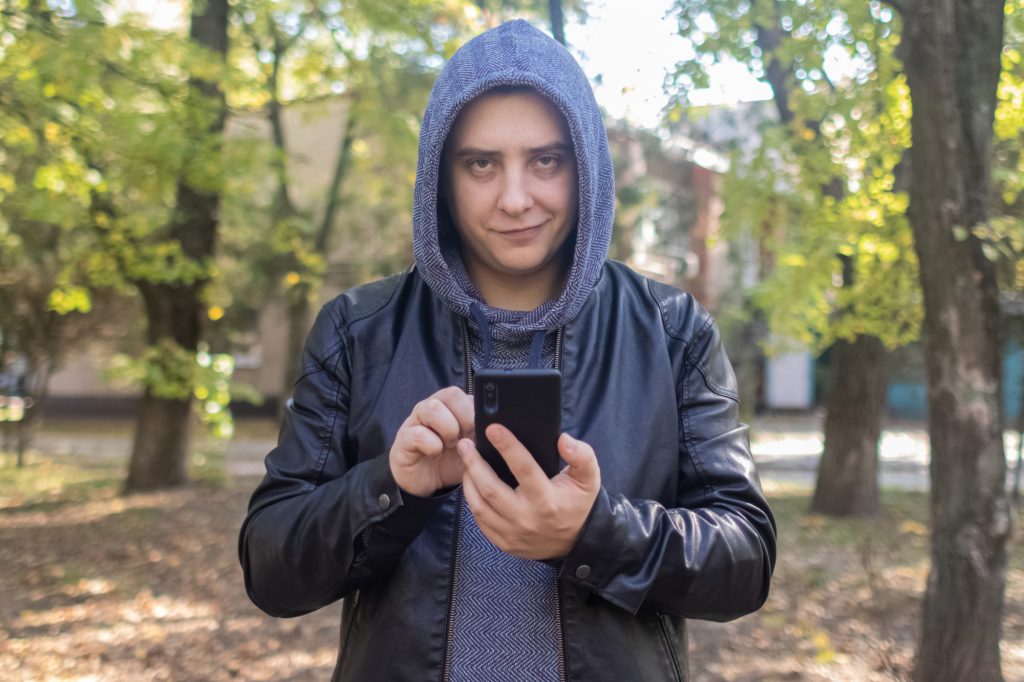 Concept. A man in a leather jacket with a hood looks at the phone and smiles like a maniac or a telephone scammer. Theft of information from a smartphone. Correspondence of a maniac on social networks