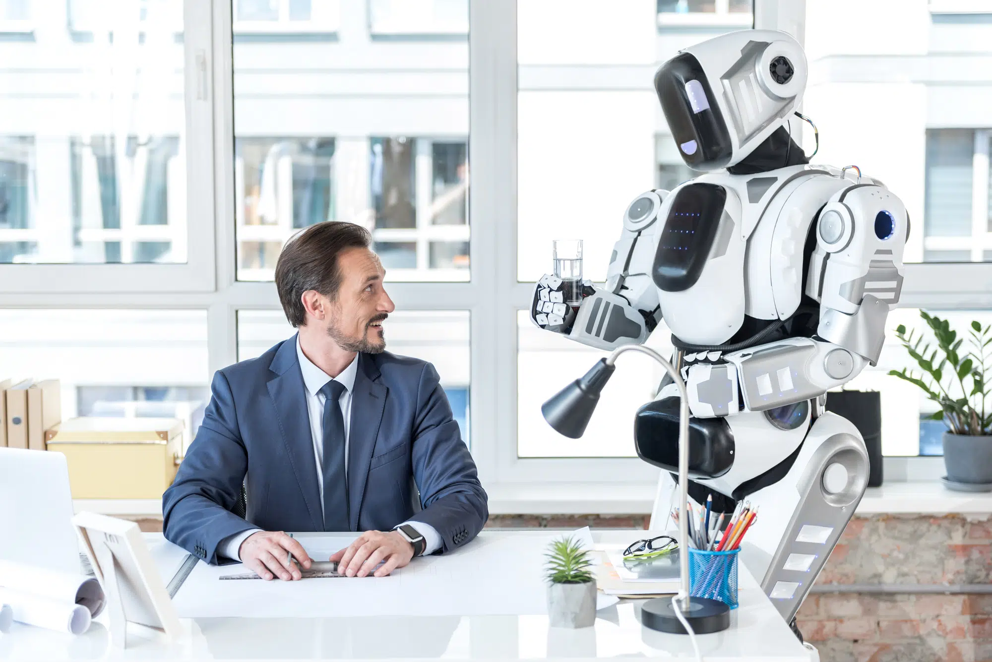 This is for you. Pleasant positive smiling man is sitting at table and working while getting glass of water from robot