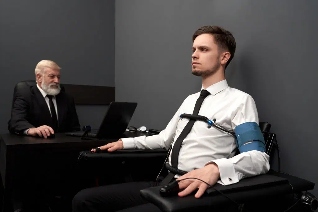 Young man passing check with lie detector, sitting on black chair. Sensors with wires on fingers of hands and chest. Elderly expert examinating man with computer polygraph and defining lie or truth.