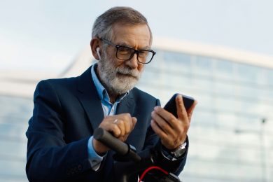 Senior gray-haired man in glasses and headphones standing at bike on street and tapping or scrolling on mobile phone . Old grandfather in airpods using smartphone and listening to music outdoor.