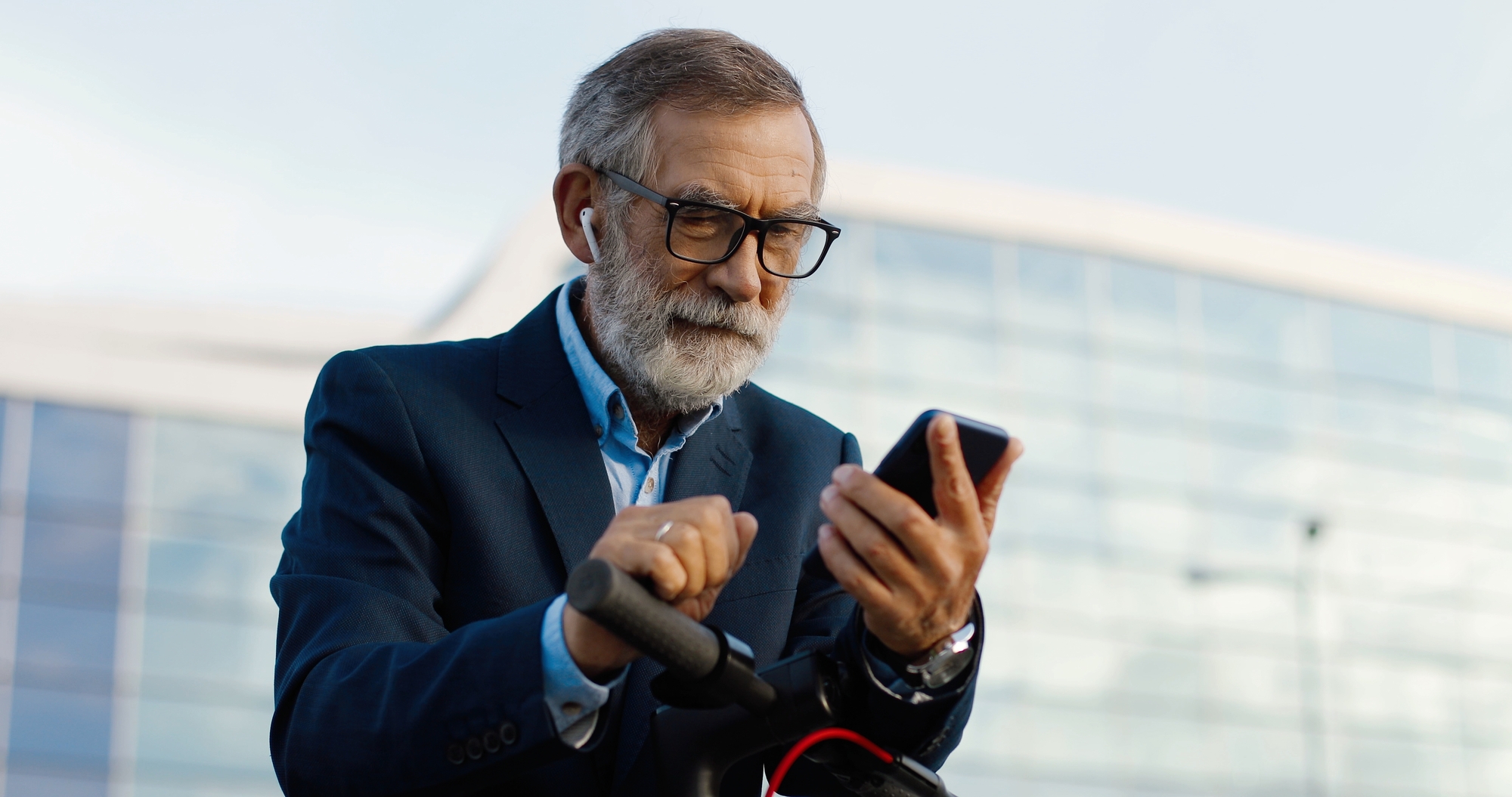 Senior gray-haired man in glasses and headphones standing at bike on street and tapping or scrolling on mobile phone . Old grandfather in airpods using smartphone and listening to music outdoor.