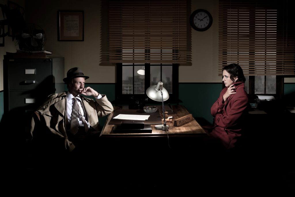 Handsome detective at office desk interviewing a young woman, 1950s film noir style.