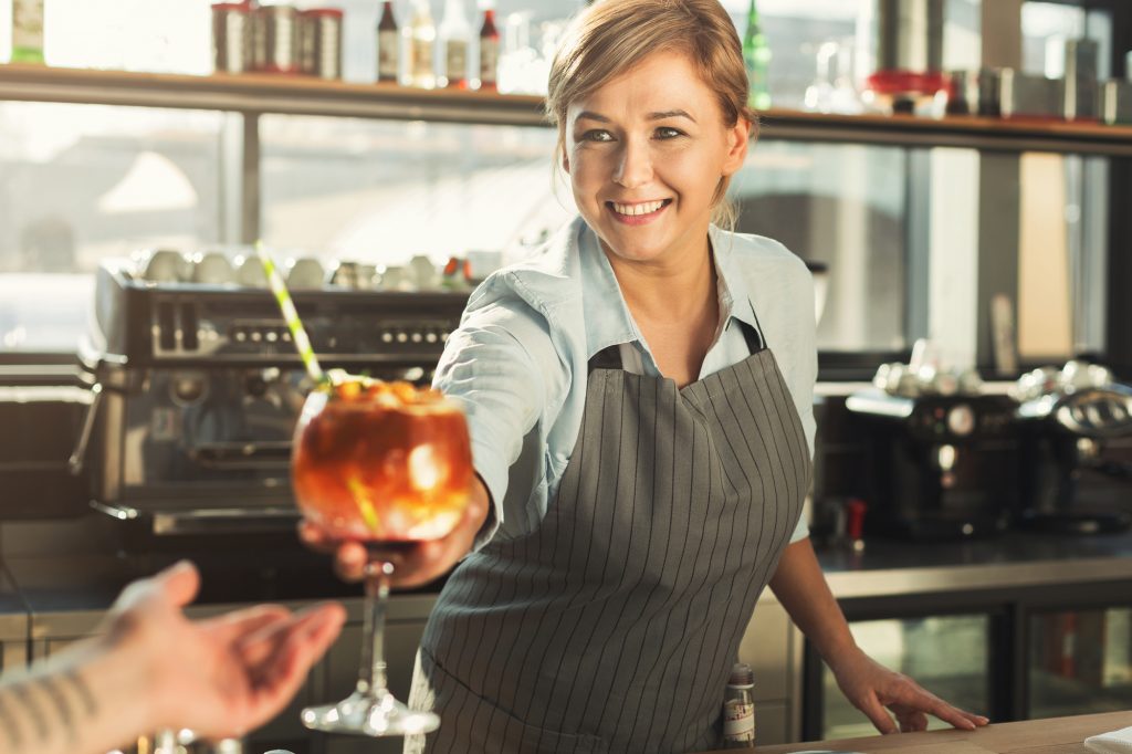 Experienced barista serving alcohol cocktail in cafe. Woman giving beverage in glass with ice cubes and decoration. Small business and professional barman concept
