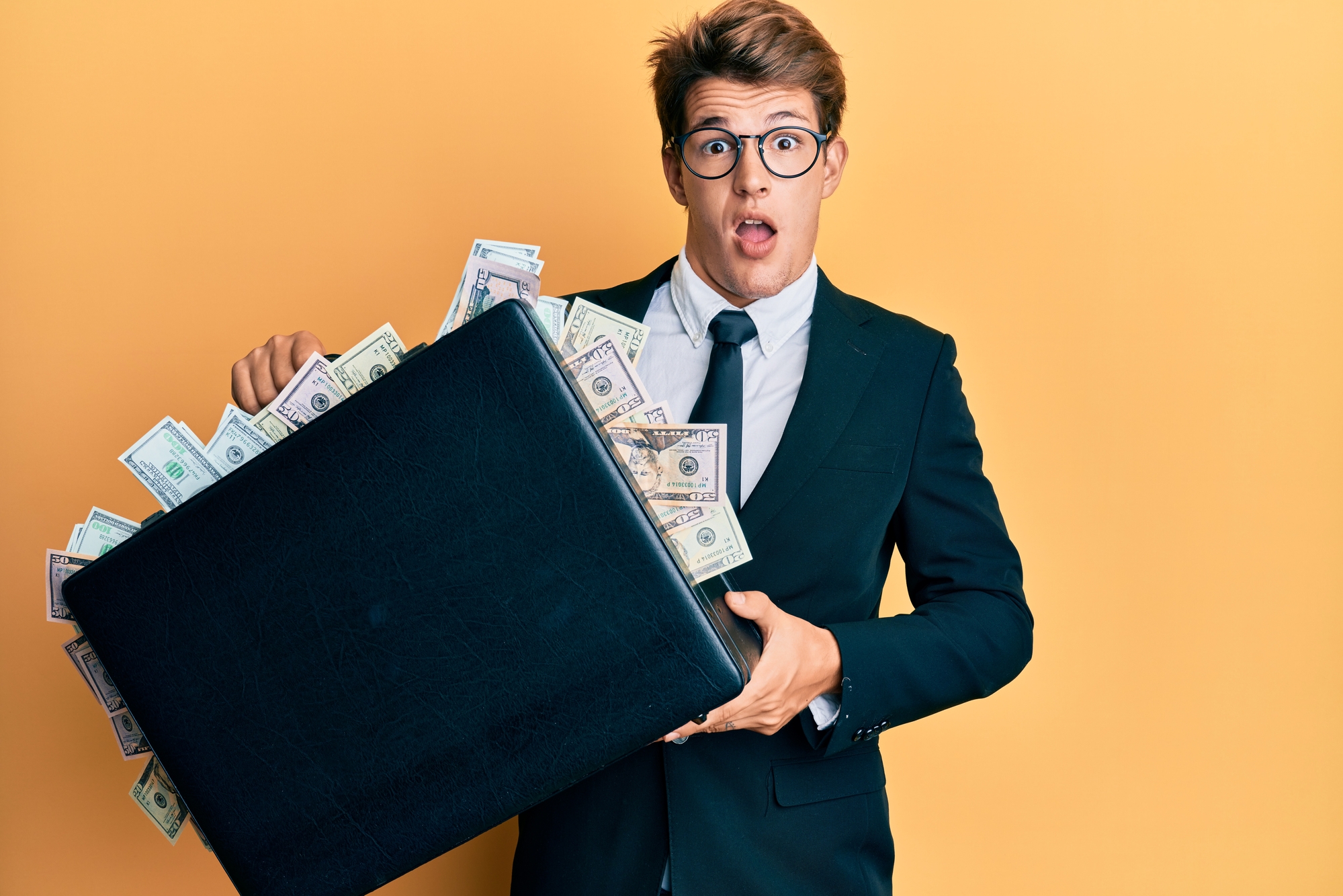 Handsome caucasian business man holding briefcase full of dollars afraid and shocked with surprise and amazed expression, fear and excited face.