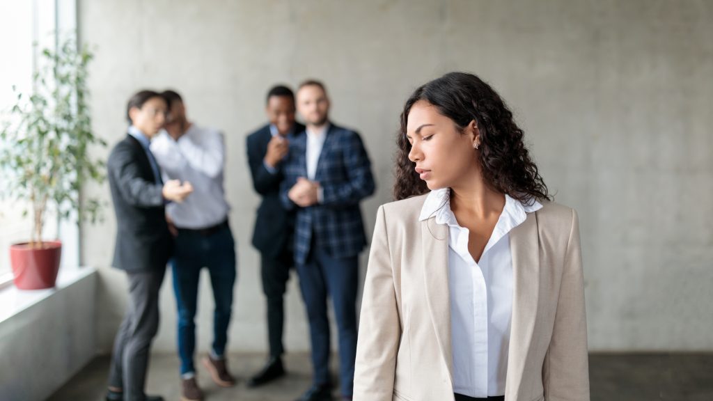 Sexism. Group Of Businessmen Whispering Behind Back Of Victimized Latin Businesswoman In Modern Office. Females Discrimination Problem, Workplace Bullying Concept. Panorama, Selective Focus