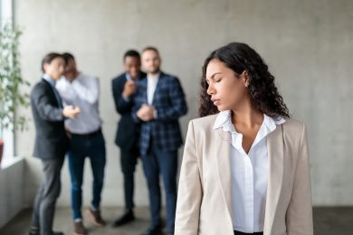 Sexism. Group Of Businessmen Whispering Behind Back Of Victimized Latin Businesswoman In Modern Office. Females Discrimination Problem, Workplace Bullying Concept. Panorama, Selective Focus