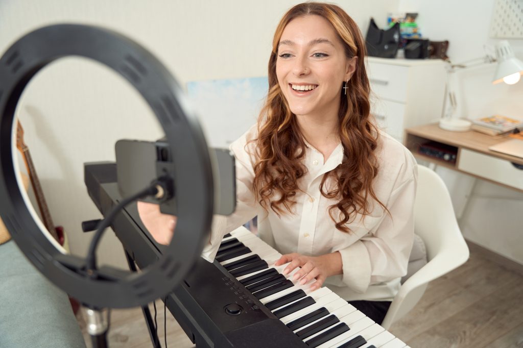 Smiling pleased young keyboardist sitting at synthesizer and adjusting settings on cellphone