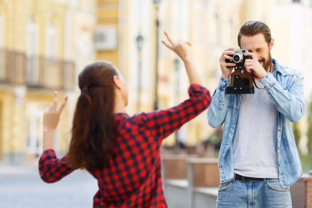 Lets make funny pictures. Pleasant bearded vivacious man holding camera and making photo of the girl posing in front of it