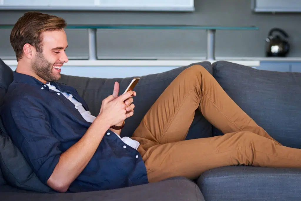 Profile image of handsome man busy smiling while texting on his mobile phone, comfortably seated on his modern couch in his living room with the sleek kitchen visible behind him.