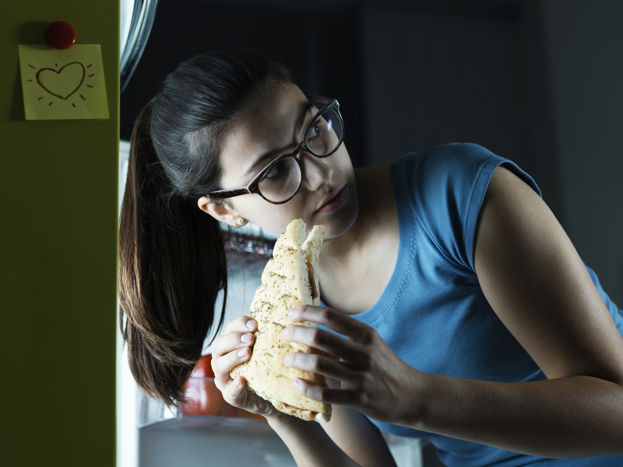 Young woman having a late night snack, she is taking a sandwich fron the fridge and eating it, diet fail concept