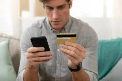 Front view close up of a young Caucasian man sitting on a sofa at home using a smartphone and holding his credit card