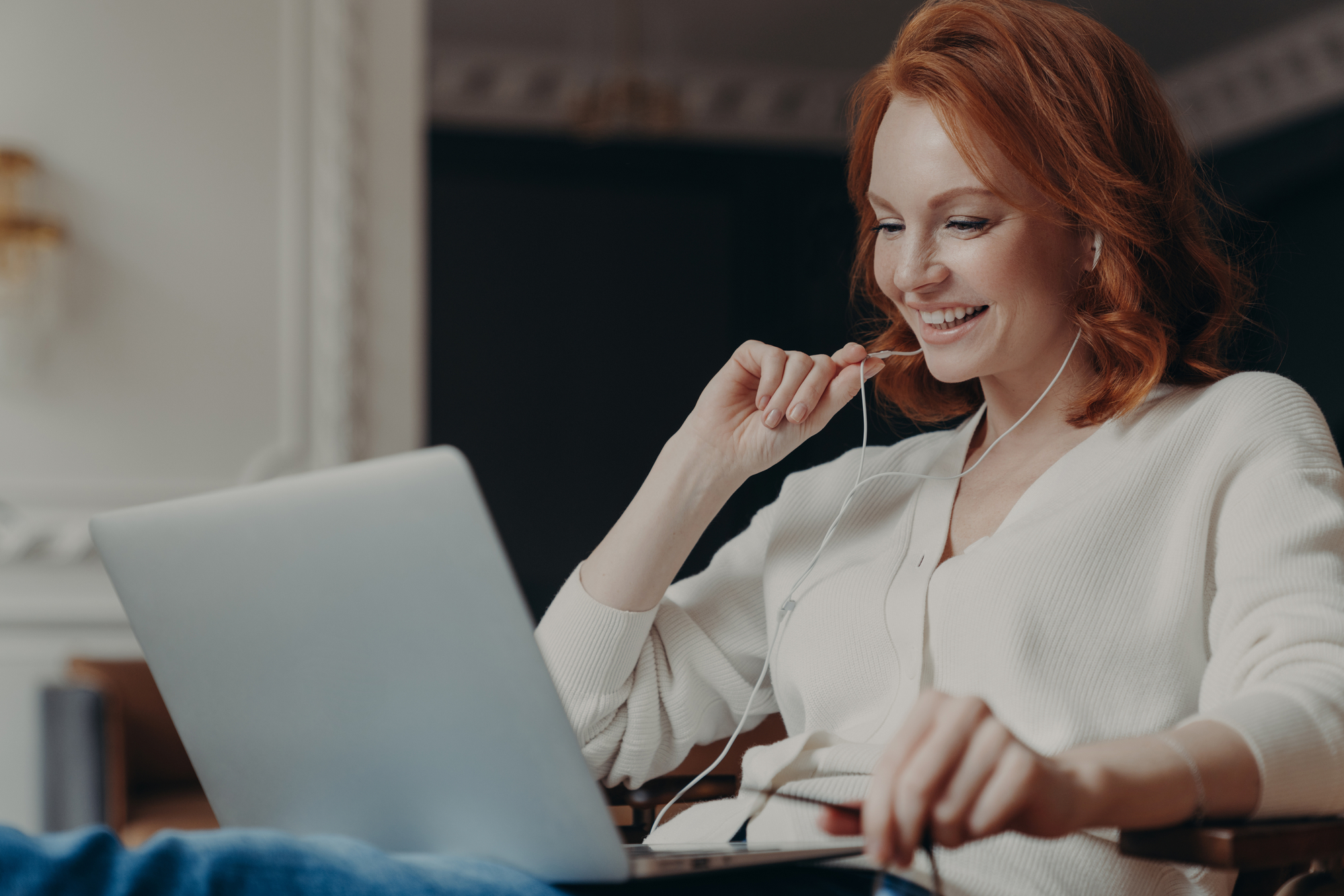 Positive redhead woman studies online course, talks with friend in application, uses earphones, enjoys video conference from favorite blogger, browses internet. Distant communication and work