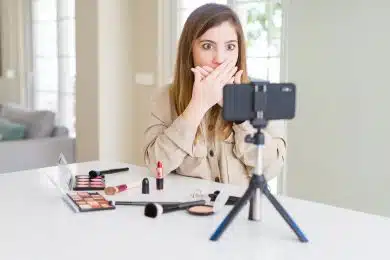 Beautiful young influencer woman recording make up video tutorial shocked covering mouth with hands for mistake. Secret concept.