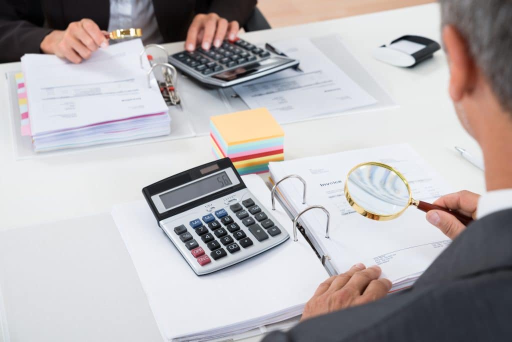 Close-up Of Businesspeople Scrutinizing Bills With Magnifying Glass