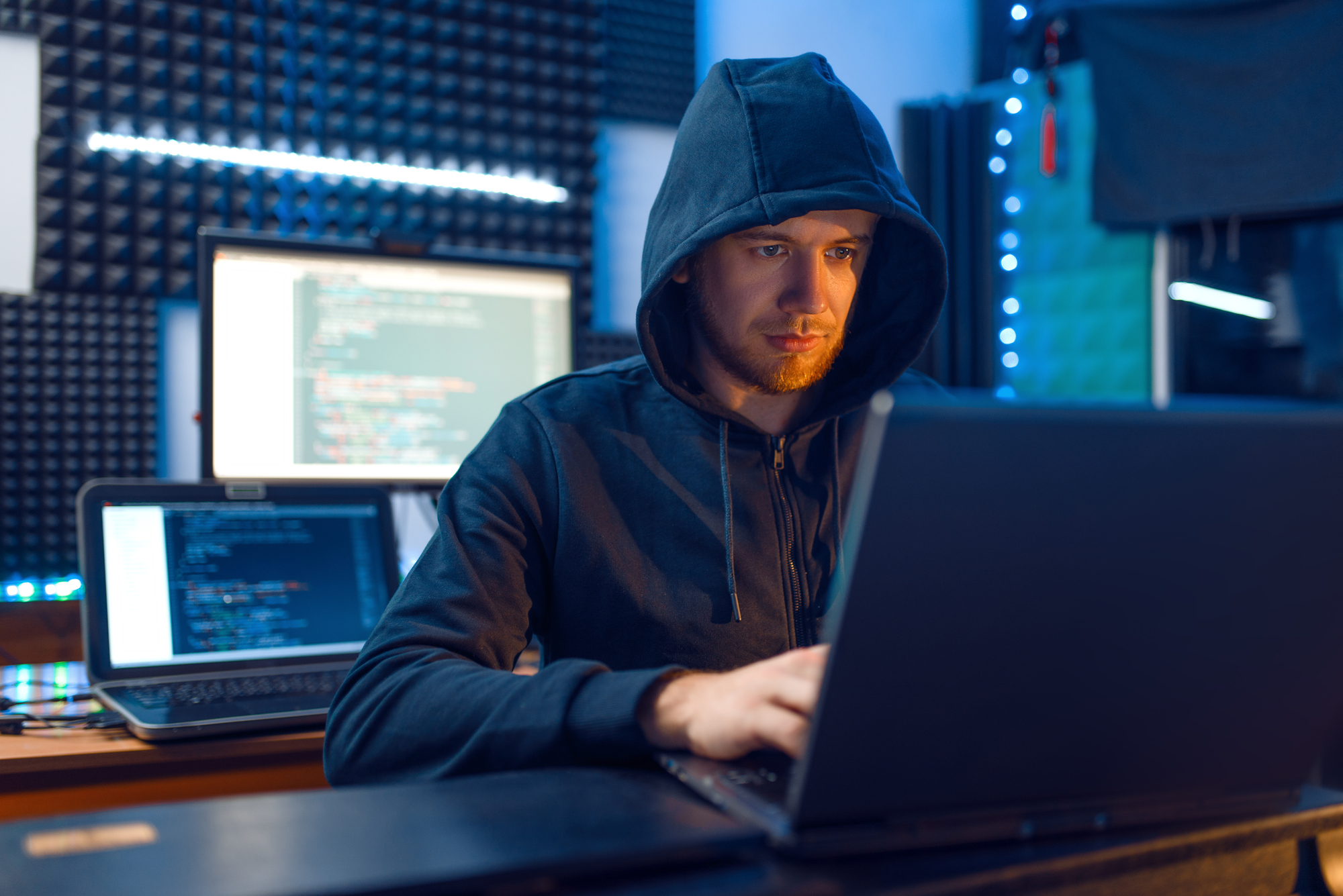 Hacker in hood at his workplace with laptop and desktop PC, password or account hacking, darknet user. Internet spy, crime lifestyle, risk job, network criminal