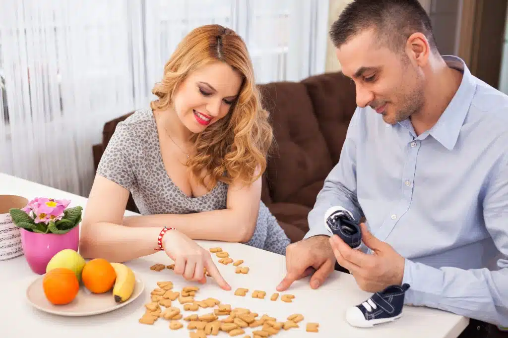 Pregnant young woman and her husband making their baby name using letters from biscuits, sitting at the kitchen table
