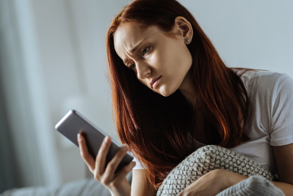 Nothing good. Depressed unhappy young woman looking at her smartphone screen and feeling unset while receiving an unpleasant message