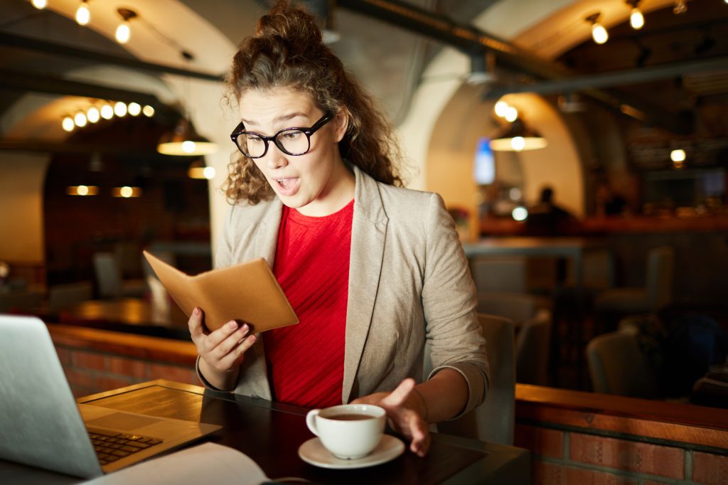 Young amazed businesswoman looking at bill with price of her order while sitting in cafe