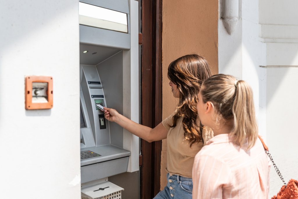 Two beautiful tourists female withdrawing the money from a bank card using ATM machine at the city center. Two travelers friend withdraw money for lunch and coffee