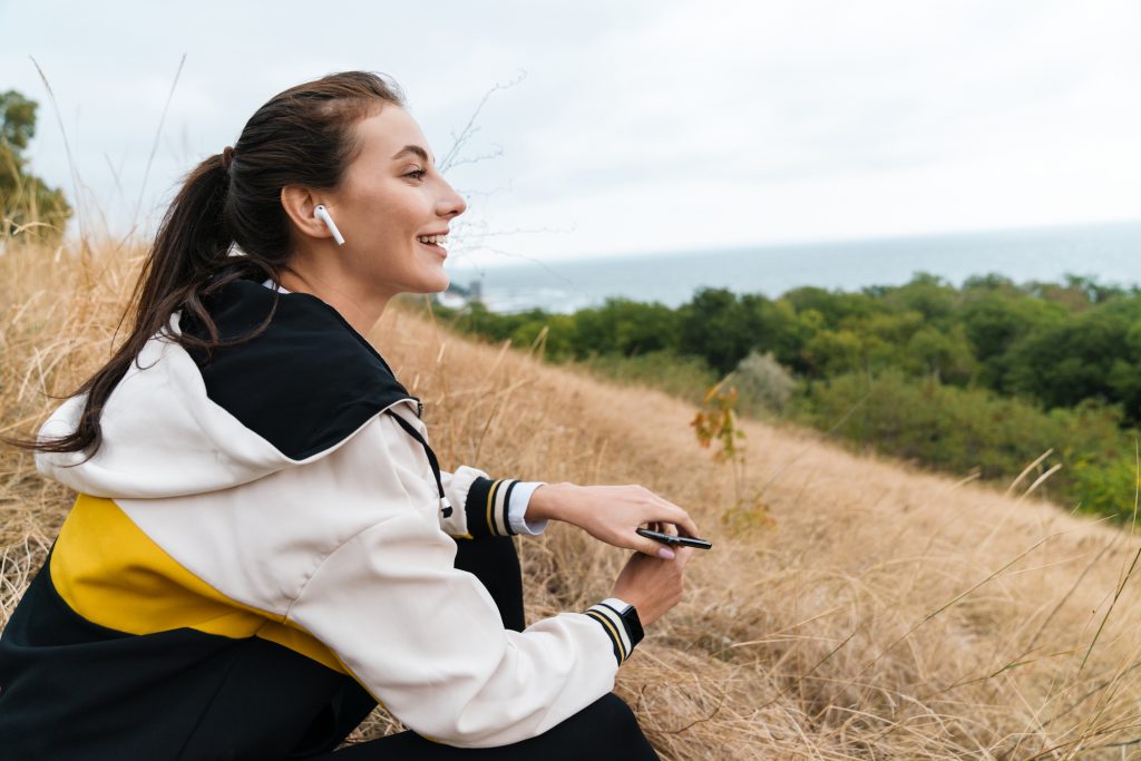 Photo of nice happy woman in sportswear using earpods and smartphone while sitting on dry grass at seashore