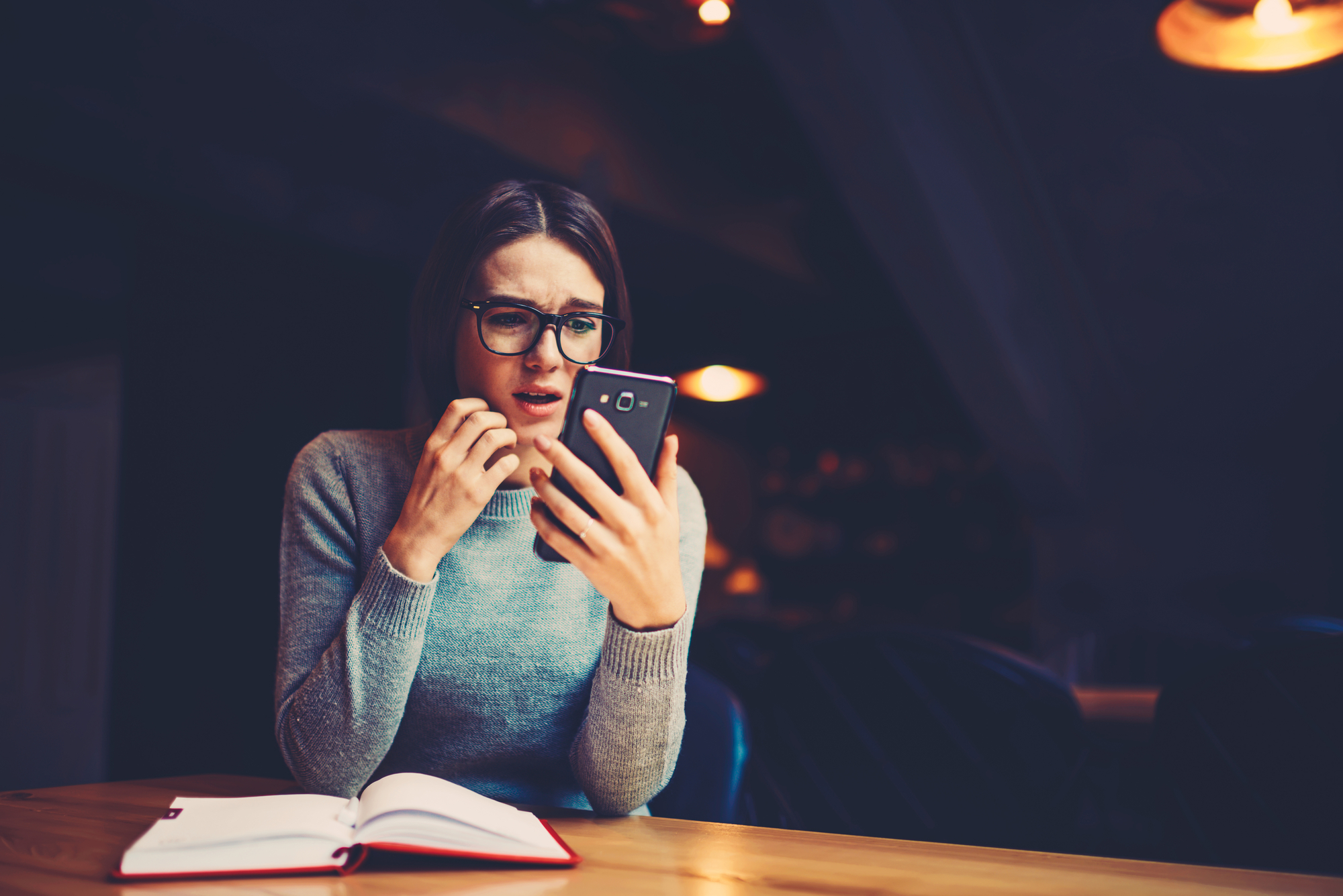 Hipster girl in eyeglasses worrying and watching online broadcasting on mobile phone while preparing for exam.Emotional female reading student's rating on university website in expecting good results