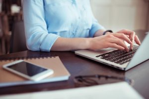жінка в блакитній сорочці працює за ноутбуком, видно тільки руки